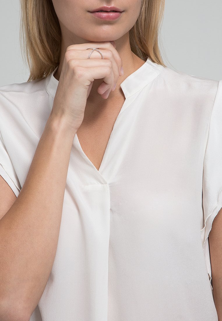 Weiße Bluse mit kurzen Ärmeln und V-Ausschnitt. Weiche, glatte Textur. Die Hand des Models ist mit einem zarten, silbernen Ring geschmückt, der wie ein V geformt ist.