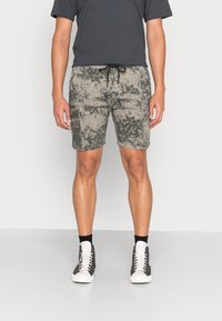 Bermudas de camuflagem em bege e cinza com bolsos laterais, cintura com cordão e um corte relaxed, combinadas com sapatilhas escuras e meias brancas.