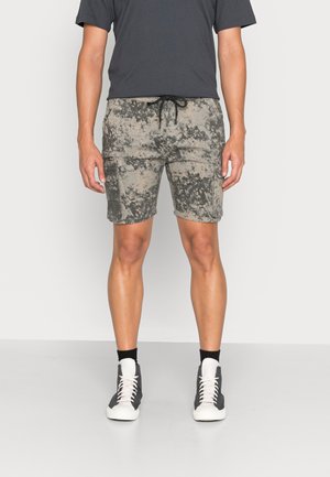 Bermudas de camuflagem em bege e cinza com bolsos laterais, cintura com cordão e um corte relaxed, combinadas com sapatilhas escuras e meias brancas.