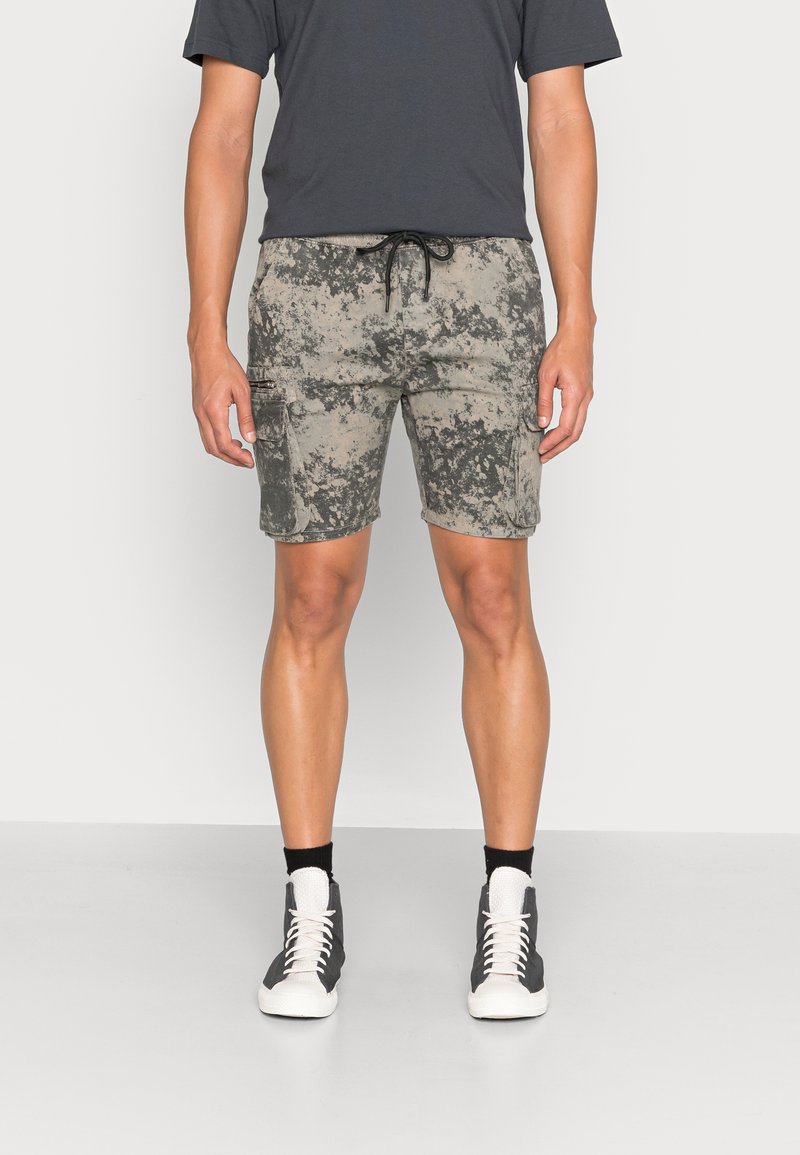 Bermudas de camuflagem em bege e cinza com bolsos laterais, cintura com cordão e um corte relaxed, combinadas com sapatilhas escuras e meias brancas.