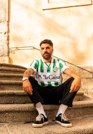 Groen-wit gestreepte voetbalshirt met logo, gecombineerd met zwarte broeken en witte sokken, zittend op stenen trappen tegen een lichte muur.