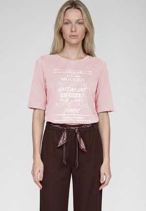 Frau trägt ein pinkes, kaffeebezaubertes T-Shirt und dunkelbraune Hose mit gemustertem Halstuch als Gürtel, steht vor schlichtem grauem Hintergrund.