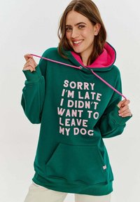 Zelená mikina s kapucí lemovanou růžovou, se spoustou bílého textu: "OMLUVTE MĚ, ŽE JSEM OPOZDIL, NECHTĚL JSEM OPUSTIT SVÉHO PSA." Obsahuje přední kapsu.