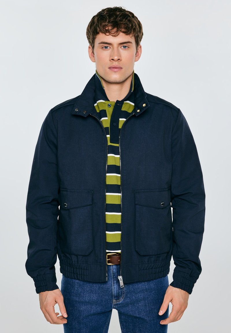 Tumesinine jakk, millel on kõrge krae, lukk, kaks esimest taskut ja elastsed varrukapitsid. Allpool paistab roheline ja tumesinine triibuline särk.