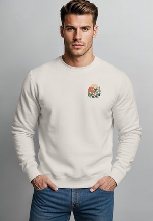 Young man with styled hair wearing a white sweatshirt with a small nature-themed graphic and blue jeans, hands in pockets.