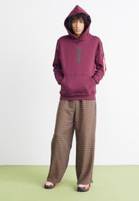 Sudadera granate con bolsillo frontal y parche de logo, combinada con pantalones anchos de cuadros en marrón, naranja y amarillo. Zapatos negros con acentos rojos.