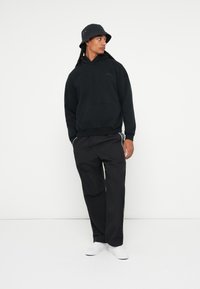 Črna kapuca z sprednjim žepom, v kombinaciji s črnimi širokimi hlačami. Dopolnjeno s črnim klobukom in belimi tenisicami.