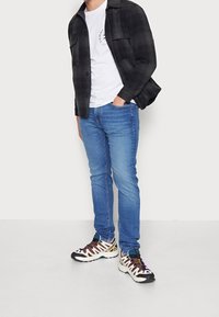 Camicia a quadri neri, t-shirt bianca, jeans skinny blu e sneakers multicolori con lacci evidenti e suole spesse.