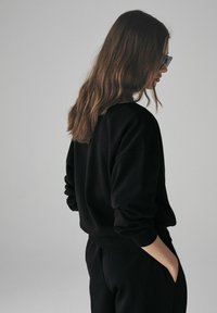 Felpa nera con maniche lunghe, polsini e orlo a coste. Il modello sta in posizione laterale, con le mani in tasca, capelli mossi e scuri. Sfondo neutro.