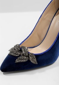 Chaussure à talon haut en velours bleu marine avec un bout pointu et un détail décoratif en forme de feuille ornée de noir sur le dessus.