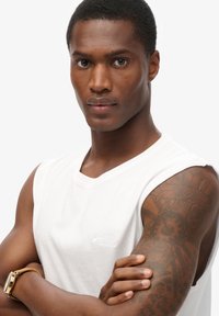 Camisa sin mangas blanca con cuello redondo, textura suave y logo bordado minimalista. Exhibición de un tatuaje marrón en el brazo.