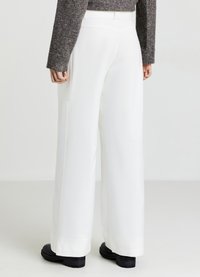 Pantaloni bianchi a gamba larga con una texture liscia, dotati di tasche laterali e una cintura pulita. Abbinati a un maglione scuro in maglia e calzature nere.