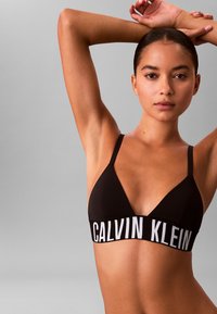Zwarte driehoekige bralette met dunne bandjes en een brede band met "CALVIN KLEIN" in het wit. Gladde textuur, minimalistisch ontwerp.