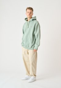 Light green hooded sweatshirt with a front pocket and drawstring, paired with beige relaxed-fit pants and white sneakers. Simple design.