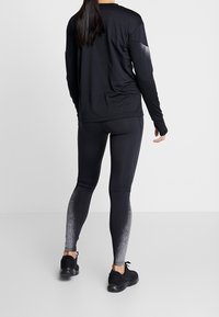 Vrouw staat rechtop, gekleed in een zwart langemouwen top en zwarte leggings met een wit gespikkelde verloop op de onderbenen, zwarte sportschoenen, tegen een effen achtergrond.