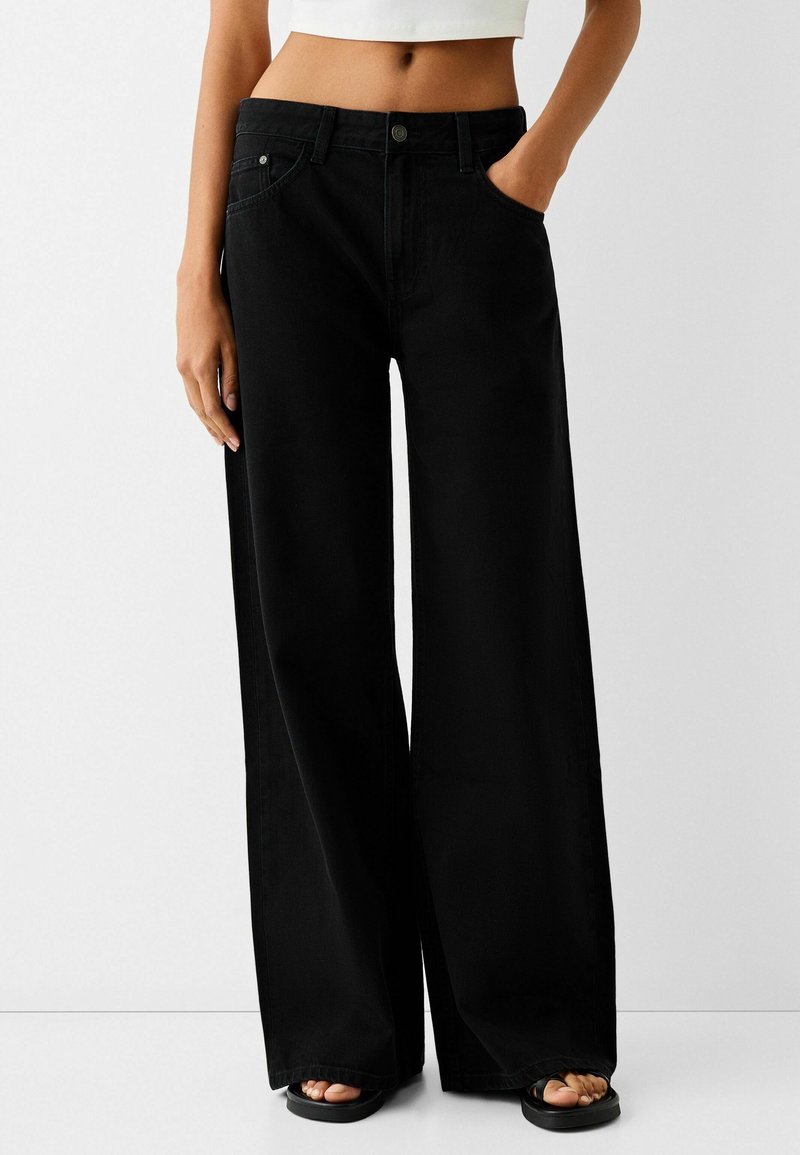 Femme portant un pantalon noir taille haute à jambes larges, main dans la poche et sandales noires à bouts ouverts, montrée de la taille aux pieds.