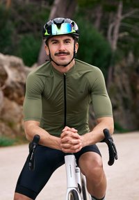 Cycliste portant un maillot ajusté vert olive et un short noir, assis sur un vélo blanc avec des poignées noires, les mains jointes devant.