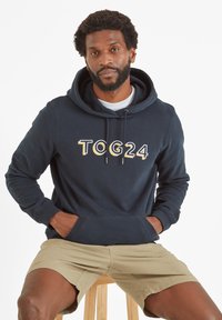 Man with beard wearing navy blue "TOG24" hoodie and beige shorts sitting on wooden stool against white background.