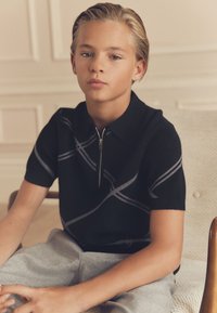Adolescent garçon aux cheveux blonds courts portant une chemise noire zippée à manches courtes et un pantalon gris, assis sur une chaise beige à l'intérieur.