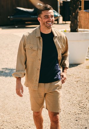 Beige linnen overhemdjasje met knopen over een navy t-shirt, gecombineerd met beige shorts. Gelaagde stof, ontspannen pasvorm, buitenomgeving.