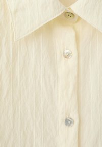 Chemise de couleur crème avec un col pointu et trois boutons visibles en nacre sur le devant.