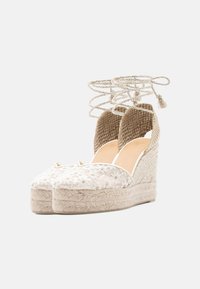Espadrilles à semelle compensée beige avec dentelle florale sur l'avant, talon tressé et liens de cheville tressés avec pompons.