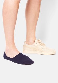 Marineblauer No-Show-Socke auf dem linken Fuß, beigefarbener Wildlederschuh auf dem rechten Fuß, der kontrastierende Materialien und Designs präsentiert.