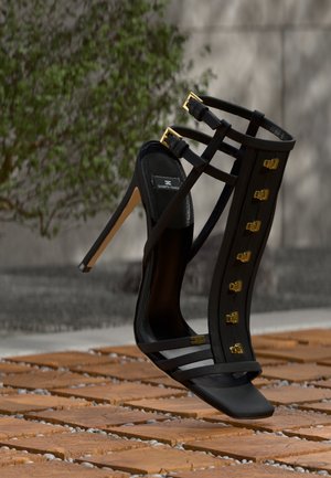 Sort højhælet sandal med guldspænder og stropper, stående på teksturerede terrakottafliser med sløret grønt løv i baggrunden.