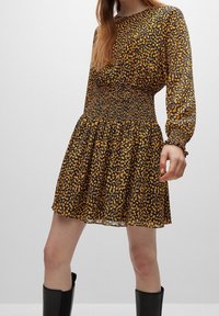 Zwarte en gele luipaardprint jurk met een gerimpelde taille, lange mouwen en een wijd uitlopende rok. Gemaakt van lichte stof met een doorschijnende textuur.