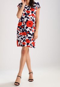 Vestido de manga corta con estampado floral sobre un fondo blanco, que presenta flores rojas y azul marino. La tela es suave y el corte es hasta la rodilla. Se acompaña de tacones negros.