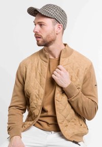 Tarnfarbene Steppjacke mit hohem Kragen, über einem einfarbigen, tarnfarbenen Pullover getragen. Das Modell trägt eine karierten Mütze und präsentiert einen lässigen, abgestimmten Look.