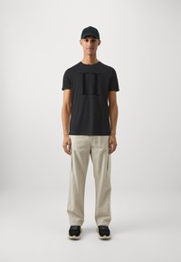 T-shirt noir à manches courtes avec un graphisme "II" en évidence, associé à un pantalon cargo beige clair et des baskets noires. Casquette décontractée sur la tête.
