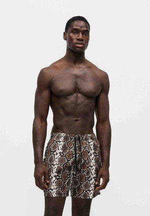 Hombre musculoso con pantalones de baño marrones y negros con estampado de serpiente parado frente a un fondo blanco liso.