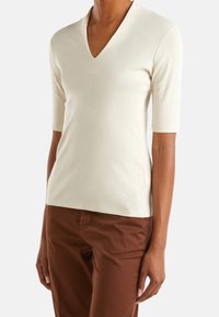 V-ringad, krämfärgad topp med korta ärmar, tillverkad av ett mjukt material, med en sidoklyfta och en liten logotyp nära fållen.