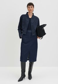 Veste en denim bleu foncé avec fermeture à bouton et deux poches avant, associée à une jupe en denim assortie avec une fente avant et des bottes noires jusqu'au genou.
