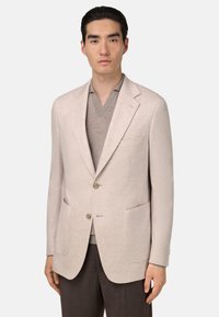 Blazer beige clair avec deux poches avant, design à boutonnière simple, revers crantés et deux boutons. Porté par-dessus un t-shirt gris à col en V.