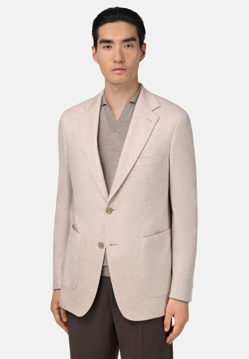 Blazer beige clair avec deux poches avant, design à boutonnière simple, revers crantés et deux boutons. Porté par-dessus un t-shirt gris à col en V.