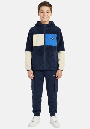 Garçon debout et souriant, portant une veste polaire zippée bleu marine et crème avec une poche poitrine bleue, un pantalon cargo bleu marine et des baskets blanches.