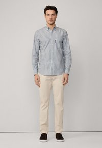 Camisa de botones a rayas en azul y blanco, de algodón, diseño ajustado. Combinada con pantalones beige y zapatos oscuros. Pose de pie contra un fondo liso.