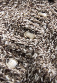 Teksturēts adīts audums brūnā, krēmkrāsā un tumšās šķipsnās; ar četriem caurspīdīgiem pogiem ar četriem caurumiem katrā; redzams detalizēts raksts.