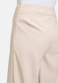 Pantalons beige clair avec une texture de tissu lisse, fermeture éclair sur le côté, taille cintrée et plis subtils pour un look structuré.