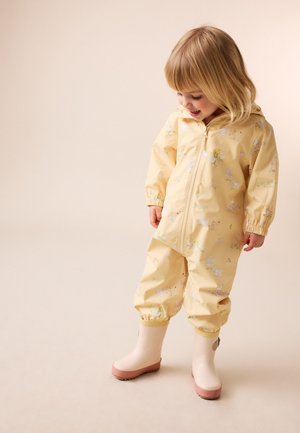 Peuter in een geel regenpak met bloemmotief en lichtroze regenlaarsjes kijkt naar beneden en glimlacht tegen een effen achtergrond.