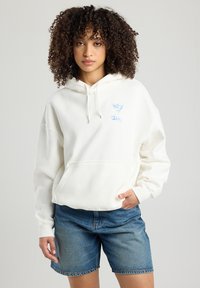 White hooded sweatshirt with a front pocket and light blue graphic design featuring a martini glass. Worn with denim shorts.