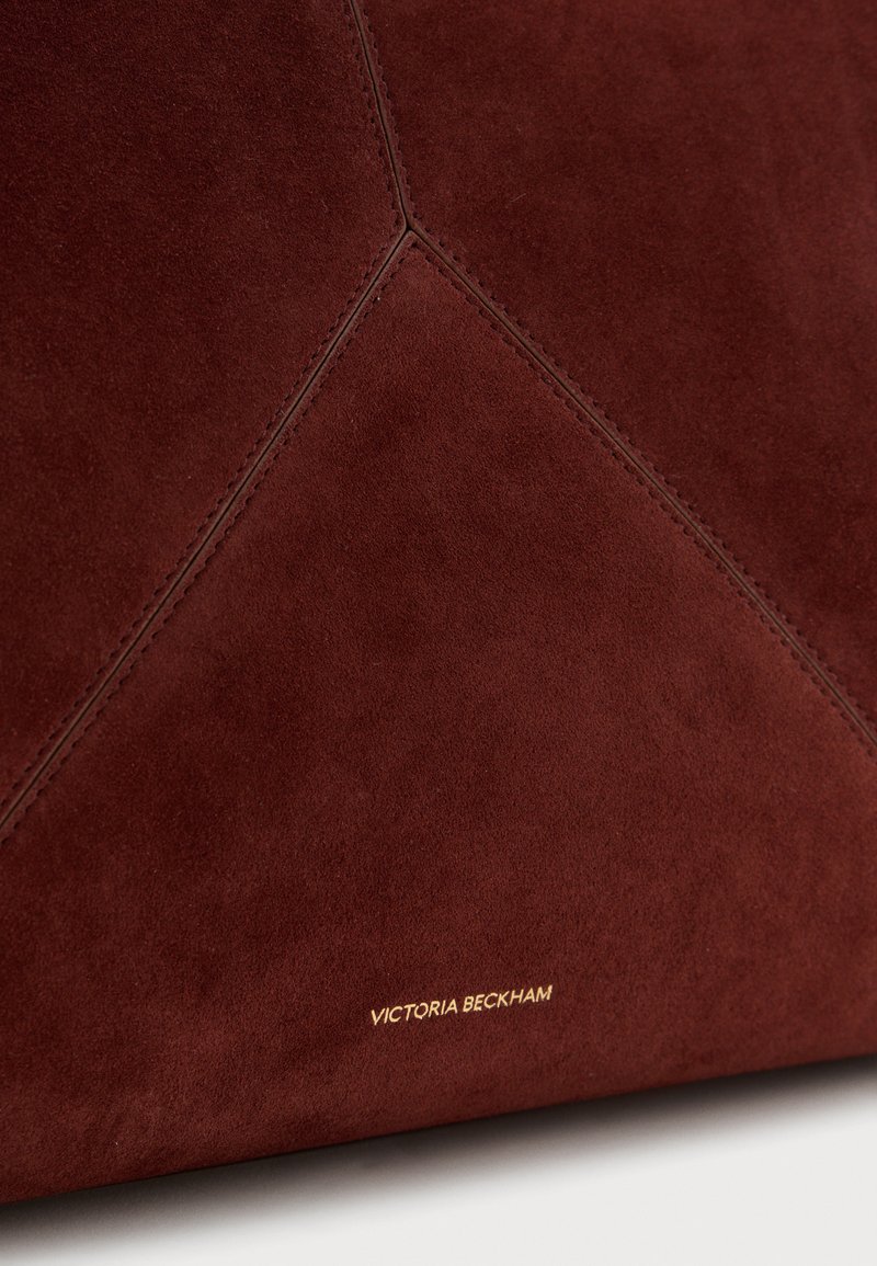 Surface en daim marron foncé avec des coutures visibles formant des motifs géométriques et "Victoria Beckham" embossé en petites lettres dorées.