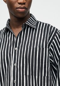 Κοντινό πλάνο ενός ατόμου που φοράει πουκάμισο με κάθετες ασπρόμαυρες ρίγες, κουμπιά, τσέπη στο στήθος και γιακά, με απλό φόντο.