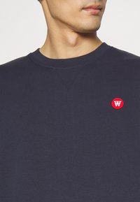Navy sweatshirt made of cotton, featuring a crew neck and a small red circular logo on the left chest area. Smooth texture, minimal design.