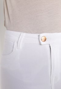 Los pantalones de mezclilla blancos presentan una textura suave, un cierre de cremallera en la parte frontal y un botón dorado, con costuras sutiles y bolsillos laterales.