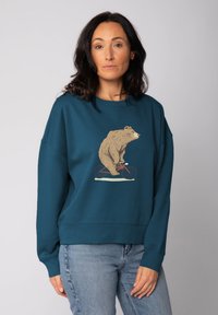 Vrouw met donker haar die een turquoise sweatshirt draagt met een afbeelding van een beer die op een rode fiets rijdt, gecombineerd met lichtblauwe jeans.
