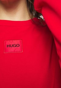 Sudadera roja hecha de un material suave, con un parche cuadrado que lleva el texto "HUGO" en negro, situado en la parte superior izquierda.