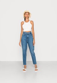 Top crop de malha branca, jeans azul de cintura alta, sandálias brancas de deslizar e um chapéu de balde bege com detalhes amarelos. Fundo simples.